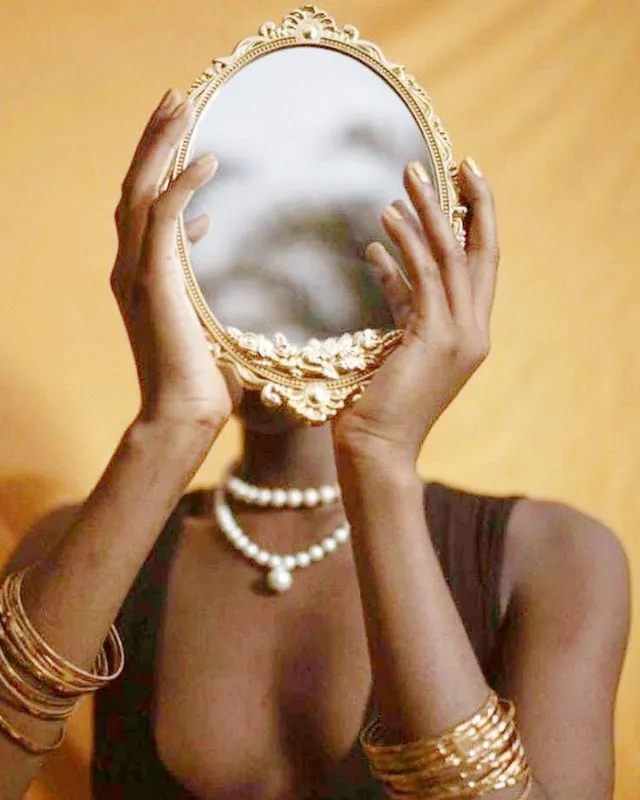 A woman holds an ornate gold hand mirror in front of her face, reflecting light against a warm gold background.